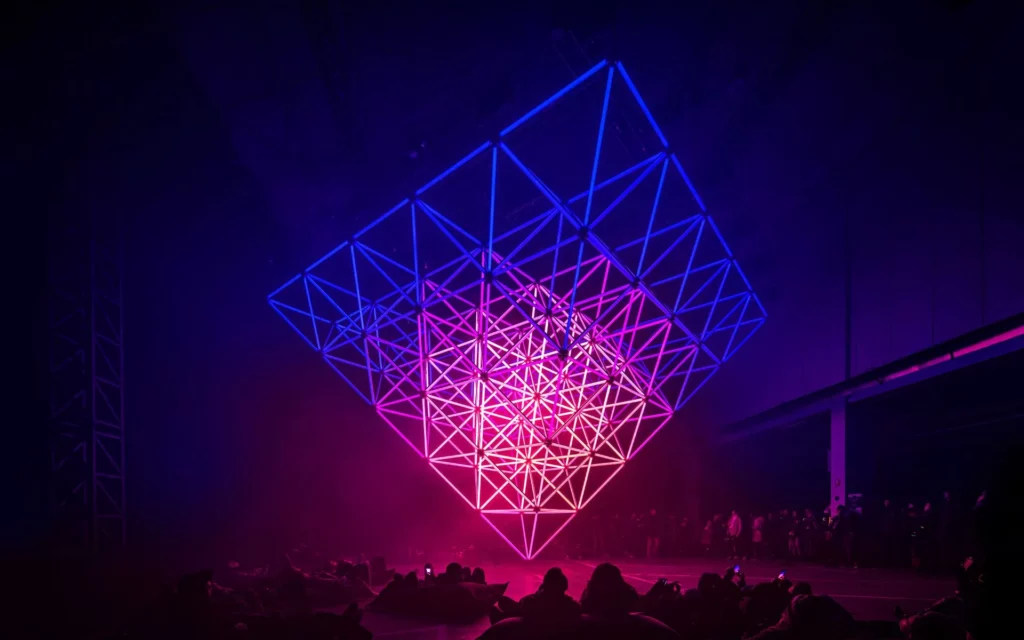 Struttura geometrica luminosa in stile diamante sospesa sopra una folla, con luci che sfumano dal blu al rosa, creando un'atmosfera futuristica e di spettacolo.
